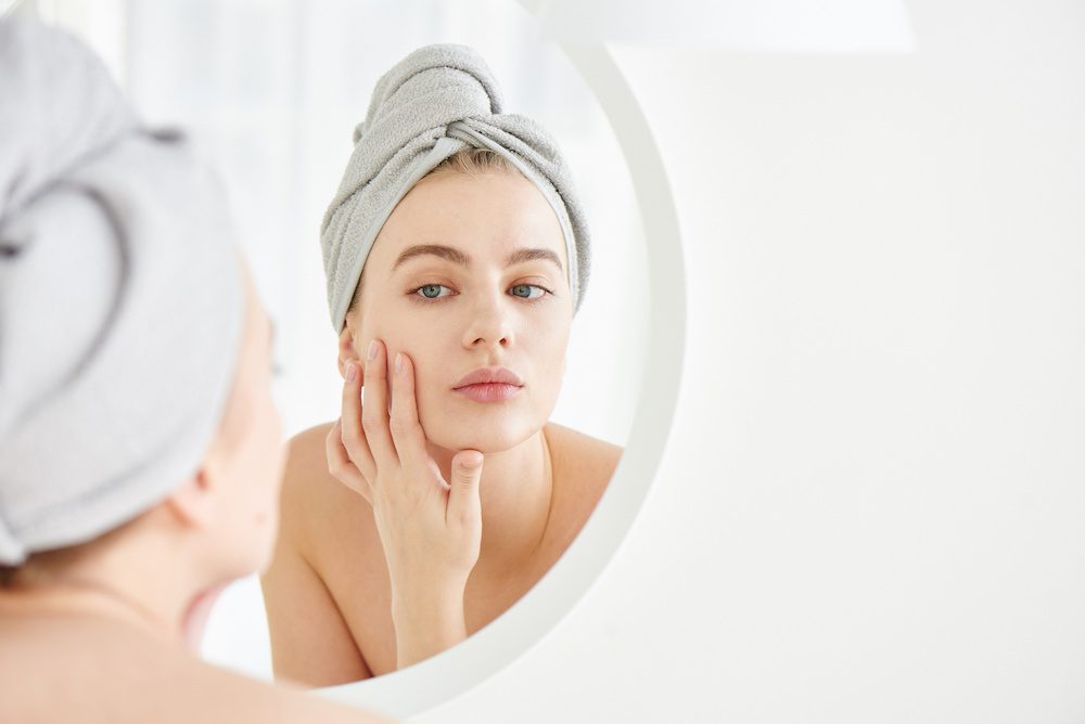 Here’s Why Young People Should Consider Botox young woman looking in the mirror at her face considering getting botox in Los Angeles.